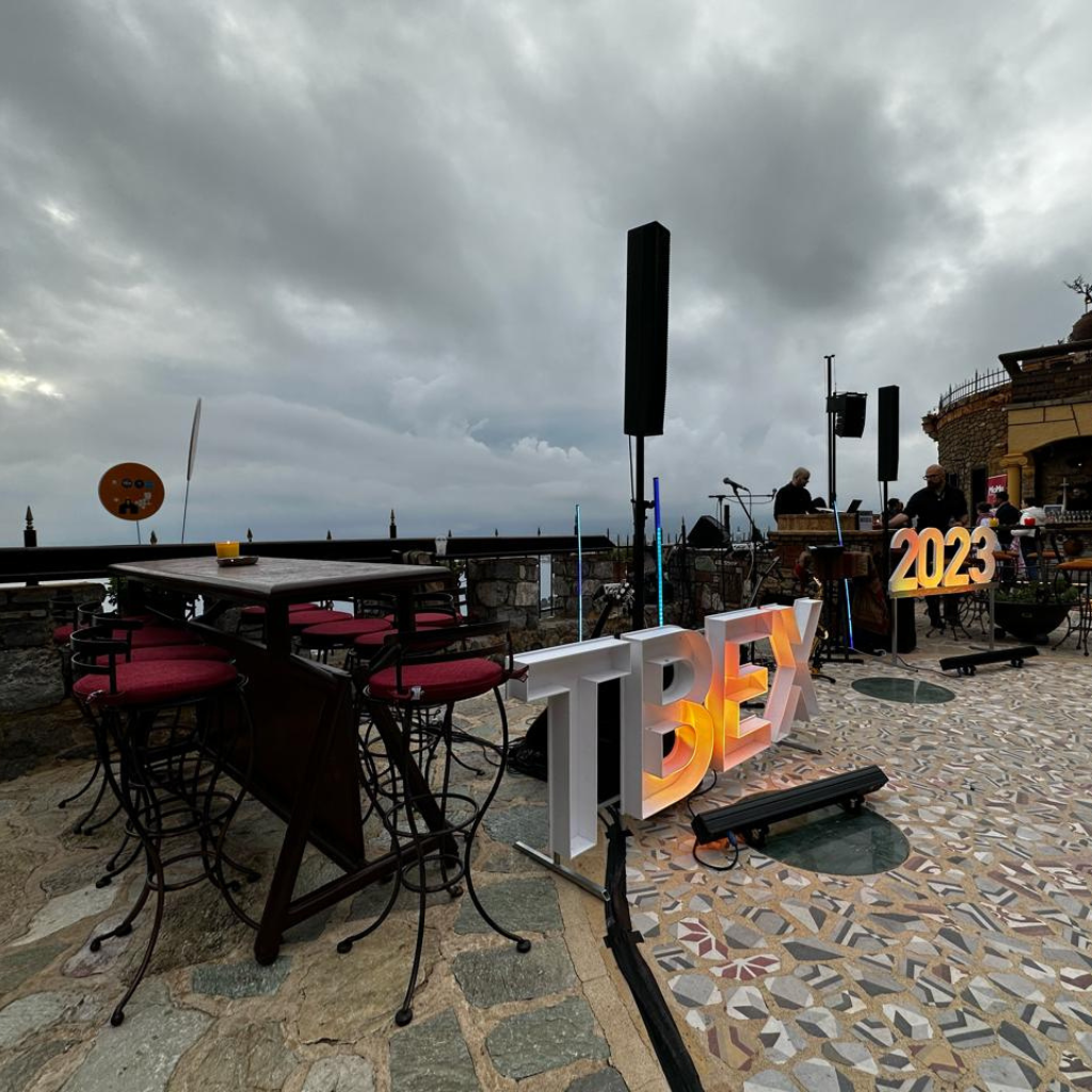 Outdoor terrace at TBEX 2023 in Kalamata, Greece, featuring a vibrant setup with illuminated 'TBEX 2023' signage, overlooking a cloudy sky and the Messinian Gulf.